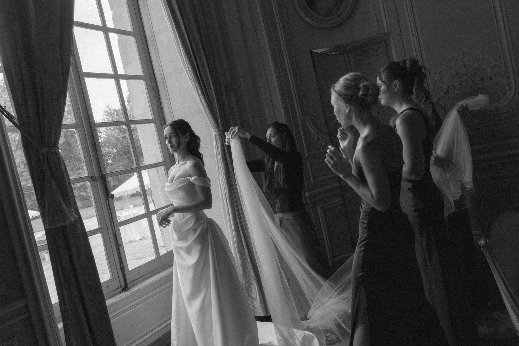 Une mariée regarde par la fenêtre d’un salon de château tandis que ses témoins déploient son voile